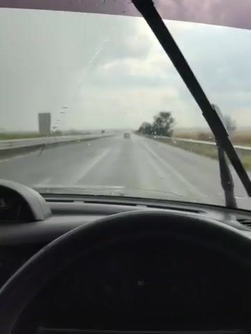 This media file is a first-person point-of-view video of driving on a highway in Orizovo, Bulgaria, during a rainstorm. The primary subject is the act of driving through the rain, with the windshield wipers actively clearing the view.

The scene is set on a two-lane highway with a median barrier. On either side of the highway, there are fields, suggesting a rural or semi-rural setting. A single tree is visible on the right side of the road, and other trees and distant vehicles can be seen through the rain. The sky is overcast and gray, indicating bad weather. The time of day is not explicitly clear, but the overcast conditions suggest it could be anytime during daylight hours.

The activity taking place is driving a vehicle. The rain is a significant element, with water droplets visible on the windshield and the road surface appearing wet and reflective. The windshield wipers are in motion, sweeping across the glass to improve visibility, which is partially obscured by the rain.

Notable details include the presence of the car's dashboard and steering wheel in the foreground, confirming the perspective. The wet conditions create a somewhat somber or perhaps peaceful atmosphere, depending on the viewer's interpretation. There is no visible text within the media file. The location context of Orizovo, Bulgaria, places the scene in that specific geographical area.