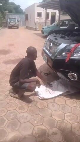 A man is squatting on a hexagonal paved surface next to the front of a black car with its hood open. He is wearing a dark t-shirt and dark shorts, and appears to be working on the car. There is a white bag with red writing on the ground in front of him. In the background, there is a building, another car, and a person walking. The lighting suggests it is daytime.