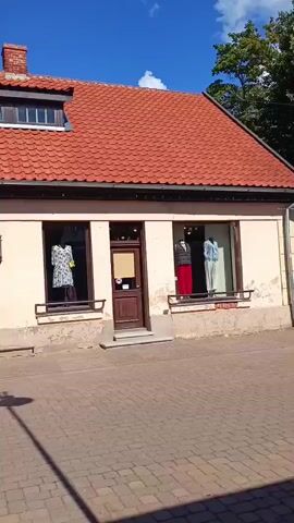 The video shows the front of a shop with mannequins displayed in the windows. The shop has a pale facade and a red tile roof. The shop windows display various clothing items on mannequins, including dresses and outfits. The pavement in front of the shop is made of interlocking bricks. A street lamp casts a shadow on the pavement. The sky is blue with some white clouds.
