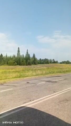 A video shows a road with white lines and some potholes. There is a grassy field with trees in the background. The sky is blue with some clouds. The video is taken from a vehicle, as indicated by the shadow in the foreground and the Infinix HOT 50 watermark. There are no people or animals visible in the video.