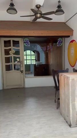 The video shows the exterior of a building with large windows and doors. The building appears to be a shop or cafe, with decorations like grape banners and fruit paintings on the walls. Inside, there are stuffed bunny toys displayed in the windows. A ceiling fan is visible, and the flooring appears to be tiled. The scene is well-lit, suggesting it's daytime.