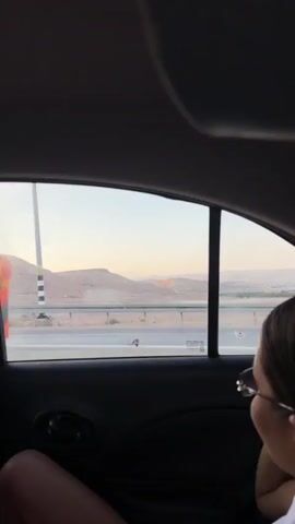 This video shows the interior of a car, viewed from the backseat. A young woman with dark hair pulled back into a bun and wearing glasses is sitting in the backseat, looking out the window. She is wearing a white shirt with a brown trim around the collar. The window shows a dry, mountainous landscape with a road in the foreground. The lighting suggests it is either sunrise or sunset. The woman's left hand is visible, resting on her lap. The car interior is dark, with the door panel and a handle visible.