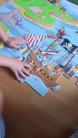 The video shows a close-up, high-angle view of a child's hands interacting with a large, colorful puzzle on a wooden floor. The puzzle depicts a pirate ship sailing on a blue ocean with several islands in the background. The ship is detailed with pirates, sails, and a Jolly Roger flag. The child's hands are positioned over the puzzle, appearing to move and adjust puzzle pieces, specifically around the ship's hull. The lighting suggests an indoor environment, possibly a living room or playroom. The overall scene is focused on the child's activity of playing with the puzzle.