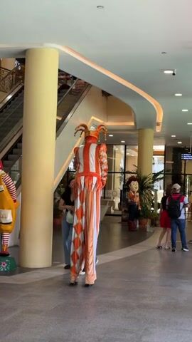 Two women are standing next to a stilt-walker dressed as a jester, posing for a photo. The jester is wearing orange and white checkered pants and a matching top with bells on the headpiece. They are in a shopping mall with an escalator in the background. A Ronald McDonald statue stands to the left, and another character statue is visible in the distance. Other shoppers are walking by.