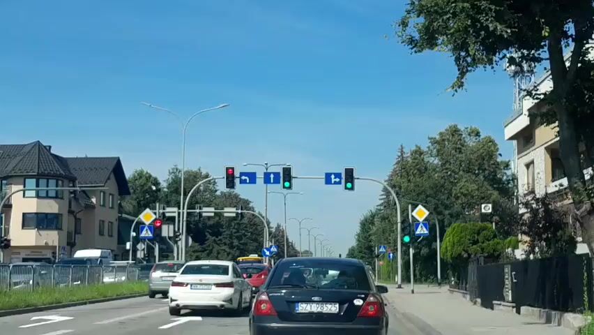 This is a first-person perspective video of driving down a street in what appears to be a European city. The sky is clear and blue. There are several cars on the road, including a white sedan, a silver sedan, and a black sedan. Traffic lights are visible, with some showing red and others showing green. There are also street signs and trees lining the road. Buildings are visible on either side of the street, some appearing to be residential and others commercial. A cell tower is visible in the distance. The video captures the car in motion, navigating through traffic.