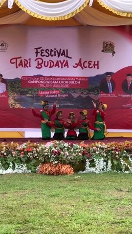 A group of five young children in traditional Acehnese dance costumes are performing on a stage. The children are dressed in red tops with green sashes and yellow headdresses. They are positioned in a line across the stage. The stage is decorated with a floral arrangement. Behind the children is a large banner that reads "FESTIVAL TARI BUDAYA ACEH". The banner also features images of landscapes and portraits of officials. The performance is taking place outdoors, with a grassy area in front of the stage. The overall scene suggests a cultural festival or performance.