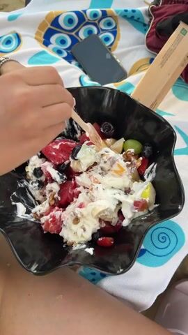 The video shows a close-up of a black bowl filled with a fruit salad mixed with yogurt and nuts. A person's hands, adorned with several bracelets, are using a wooden fork to scoop a portion of the salad. The person is wearing a bright green bikini with blue trim. In the background, a beach towel with a blue and white turtle and evil eye design is visible, along with a dark-colored smartphone. Further in the background, other beachgoers are lounging on chairs. The person then brings the fork to their mouth and eats the salad. The video appears to be taken at a beach or poolside location.