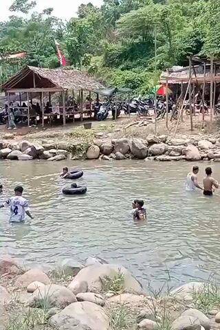 A wide shot shows a group of people swimming and tubing in a shallow, rocky river. On the riverbank, there are several wooden shacks with thatched roofs, some with people sitting inside. Motorcycles are parked behind the shacks. Lush green trees and vegetation cover the hills in the background. The water is murky brown.
