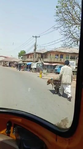 The video shows a view from inside a vehicle, likely a tuk-tuk or similar three-wheeled vehicle, driving along a paved road in what appears to be a small town or village. Several people are walking and some are standing near makeshift stalls along the side of the road.  The buildings are mostly single-story structures, with some appearing to be of older construction. Overhead power lines crisscross above the street. The sky is clear and the overall lighting suggests it is a bright, sunny day. The vehicle is moving forward, providing a moving perspective of the street scene.