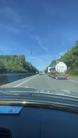 The video is filmed from the dashboard of a car driving on a highway. The car is in the right lane of a multi-lane highway. To the right of the car, there is a large tanker truck with "LPG" written on it. The highway is surrounded by trees on both sides, indicating a wooded area. The sky is blue with some wispy clouds. There are other vehicles visible in the distance on the highway. The camera appears to be slightly tilted, and there is a sticker on the lower left of the windshield that is partially visible.