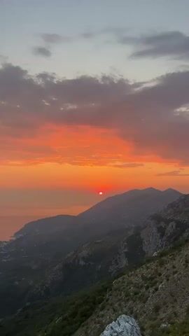 A time-lapse video shows a sunset over a mountainous coastal landscape. The sun, a vibrant orange disc, descends towards the horizon, casting a warm glow across the sky and illuminating a line of clouds with fiery hues of orange and red. The mountains in the foreground are silhouetted against the colorful sky, their rugged slopes covered in dense green vegetation. The ocean can be seen on the left side of the frame, reflecting the sunset colors. The video captures the changing light as the sun sinks lower, transitioning from a bright orange to a deeper red.