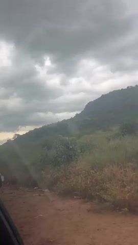 The video captures a landscape scene from a moving vehicle, likely a car given the partial view of the interior on the left. The terrain is hilly and appears to be rural, with a dirt road visible in the foreground. Lush green vegetation, including tall grasses and trees, covers the hillsides, leading up to a rocky, forested mountain in the background. The sky is overcast with heavy, dark gray clouds, suggesting impending rain or a stormy day. The camera is pointed forward and slightly upward, showing the progression of the landscape as the vehicle moves.