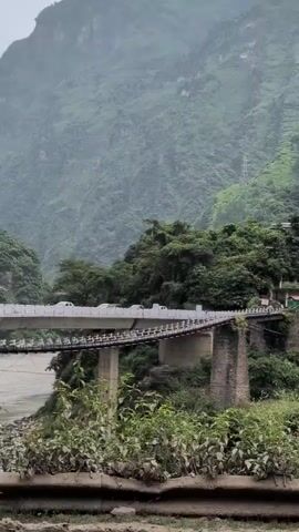 The video shows a mountainous landscape with lush green vegetation. A bridge constructed of stone pillars supports a road, with vehicles passing over it. Several buildings are scattered across the hillside, nestled amongst the trees. In the distance, a waterfall cascades down a steep slope. The sky appears overcast, suggesting a cloudy or possibly rainy day. The overall impression is one of a remote, natural setting with a small human presence.