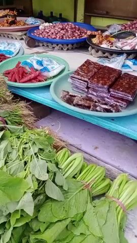 The video shows a street market stall with various food items. In the foreground, large bundles of leafy green vegetables are displayed, some with exposed roots. Behind these, there are several platters and bowls containing different products. One platter holds a pile of red chilies. Another contains what appears to be dark, rectangular blocks of food, possibly processed or dried meat or fish. There are also bowls of small red onions and a platter with what looks like dried fish or shrimp. In the background, a person wearing a pink shirt is partially visible, seemingly attending to the stall. The overall impression is of a vibrant, local market setting.