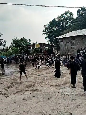 This is a video of a cultural performance or competition taking place outdoors in what appears to be a village or rural area in Gentungan, Indonesia. The setting includes a sandy or dirt ground, with some temporary structures and trees in the background. There are numerous spectators gathered to watch the event.

The main activity involves several individuals, mostly young men, engaged in what seems to be a martial arts demonstration or a traditional dance performance. Some participants are dressed in black uniforms, while others wear casual clothing. There are musical instruments and a sound system set up, suggesting a musical accompaniment to the performance. In the foreground, a person in a black outfit with a white bandana is holding a whip or a similar implement, and another person is crouched down. A crowd of onlookers is watching from the sides and behind the performers, with some standing and others seated.

The time of day appears to be daytime, likely late morning or afternoon, given the overcast sky and the general lighting conditions. The weather seems to be cloudy, and there are no obvious signs of rain. The location cues, such as the style of the buildings and the vegetation, are consistent with a tropical or subtropical environment.

There is a yellow banner with text visible in the background. While the text is not entirely legible, it appears to be a sign or advertisement for the event. The overall atmosphere seems lively and engaged, with the spectators showing interest in the performance. The participants are focused and appear to be executing their moves with precision and energy.