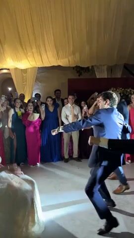 The video captures a lively wedding reception. A bride in a white gown sits on a chair while two groomsmen dance enthusiastically in front of her. They are dressed in blue and black suits. Surrounding them is a crowd of guests, many holding up their phones to record the moment. The women are dressed in a variety of bright dresses. The venue has a large, decorated ceiling and a red door in the background, contributing to the celebratory atmosphere.