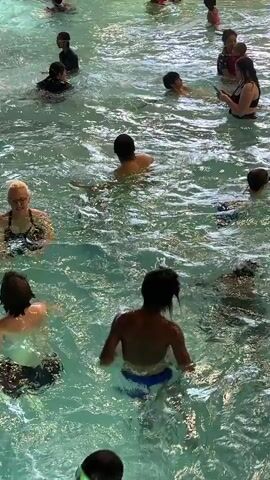 The video shows a crowded indoor water park. Several people, including children and adults, are in the water. Some individuals are swimming or playing, while others are standing or sitting in the pool. The water appears to be shallow in some areas and deeper in others. The lighting suggests an indoor environment, likely a water park with artificial lighting. The overall atmosphere is one of recreation and enjoyment.