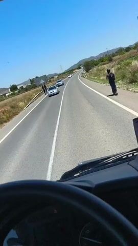 Here's a description of the video:

The video is shot from the perspective of someone driving a truck on a two-lane road. The sky is clear and blue. There is a silver car driving in the same direction as the truck, a few cars up ahead. On the right shoulder of the road, two people wearing dark clothing stand on the edge of the road, with one of them standing a bit further up the road and waving their arms. There is grassy vegetation on the right side of the road and sparse greenery on the left side, along with a few buildings in the distance. In the far distance, a large truck is visible.