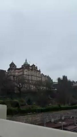 The camera pans across a view of Edinburgh, Scotland, on a cloudy day. In the foreground are train tracks extending from left to right, passing under bridges and into tunnels. The middle ground features a hillside with bare trees and low-lying greenery. Several historic buildings are visible atop the hill, including a large, domed structure and a church spire. The background is a gray sky filled with clouds. No people are visible in the video.