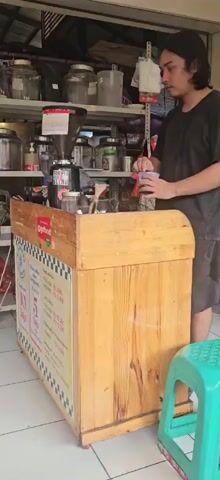 A young man with long dark hair tied back and wearing a black t-shirt and brown shorts is standing behind a wooden coffee cart. He is holding a clear plastic cup and appears to be preparing a drink. In front of him on the cart is a coffee grinder with a black base and a large clear hopper filled with coffee beans. He is operating the grinder with his right hand, pressing a button. To the left of the grinder, there are glass jars, bottles, and other items on shelves. The cart has a checkered border and a sign on its side with text and images. A green plastic stool is visible to the right of the frame. The environment appears to be an outdoor or semi-outdoor space, possibly a market stall or a shop.