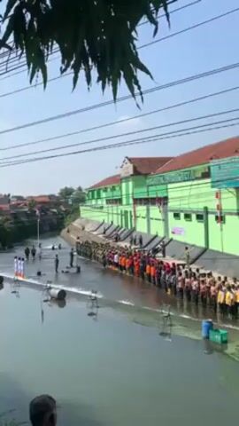 The video shows a wide shot of a river with a long line of people standing in the water along the edge. In the foreground, a few people are standing near small white, column-shaped markers. A large green building sits directly behind the line of people, with the name "SMK TRISAKTI CIKARANG" on its facade. A few individuals are standing on the riverbank in front of the building. The people in the water are wearing a variety of uniforms and outfits, including orange jumpsuits, yellow vests, and brown uniforms. A single Indonesian flag is visible on the opposite bank of the river. Power lines are strung across the sky above the scene. A tree branch partially obscures the upper left corner of the frame. The overall scene suggests a formal event or ceremony taking place in the river.