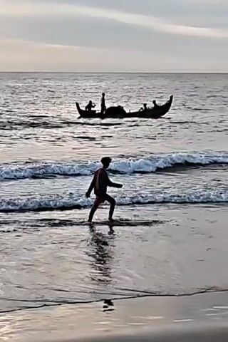 A person walks into the ocean as a boat with people on it sails further out to sea. The person is silhouetted against the water and the sky, and the boat is also in silhouette. The waves are small, and the water is calm. The sky is overcast, and the light is dim. The beach is dark sand.