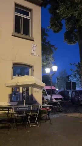 A sticker of a cartoon cat drinking from a cup is affixed to the side of a building. In the foreground, tables and chairs from an outdoor cafe are visible. A white umbrella is propped open over the tables. A street lamp with two lights illuminates the scene. A white van and other vehicles are parked in the background. Trees with dark green foliage are seen against a dark blue night sky.
