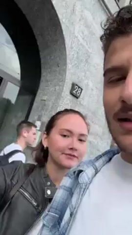 A man with curly hair and a mustache is taking a video of himself while standing outside a building. The building's exterior is a light gray color and has the number 28 on the wall. The man is wearing a white t-shirt with a light blue plaid shirt over it. He is also wearing a gold ring on his finger. Behind him and slightly to the left is a woman with her hair pulled back. She is wearing a black jacket and is partially obscured by the man. He is speaking and gesturing with his hand, and he says "I can't delete it" multiple times. The background includes the building's entrance with a glass door and part of a city street. The camera movement suggests it is hand-held and slightly shaky.