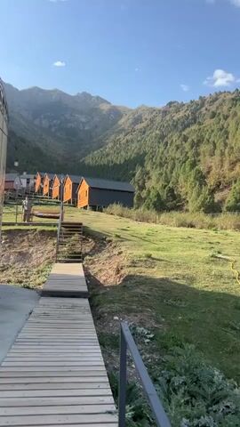 A long wooden walkway extends into the frame, leading towards a set of stairs that ascend a grassy incline. At the top of the stairs, several small, orange cabins with dark roofs are visible, lined up along the side of a hill. Beyond the cabins, a lush, green mountainside rises steeply, covered in dense trees. The sky above is a clear, bright blue with a few wispy clouds. Sunlight casts long shadows across the landscape, indicating it is either early morning or late afternoon.