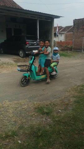Three young people are posed on a light blue electric scooter. The scooter is parked on a dirt path next to a building. Two of the people, appearing to be children, are sitting on the scooter. The third person, an older child or teenager, is straddling the scooter and leaning over the seated individuals. All three are smiling and looking at the camera. The background shows a house with a dark garage door, some greenery, and a cloudy sky. The video is oriented vertically.