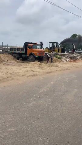 A video captures a busy construction or material handling site. In the foreground, a large orange truck is parked on a dirt surface next to a pile of sand. Several people are visible, some interacting with the sand and possibly loading the truck. To the right, a yellow bulldozer is operating, emitting a significant amount of black exhaust smoke. Piles of bagged materials, possibly cement, are stacked behind the bulldozer. Further back, a blue dump truck is being loaded with sand, with a person on top, seemingly using a shovel or tool. The background shows a body of water and trees under a cloudy sky. The overall scene depicts active, manual labor and heavy machinery operating in an outdoor industrial setting.