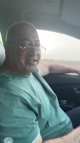 A middle-aged man with glasses and a shaved head is sitting in the driver's seat of a car. He is wearing a green t-shirt. His right arm is resting on the open window frame. The man is looking to his right and appears to be speaking. The background outside the car is blurred but suggests a rural or open landscape with a pale sky. The interior of the car is visible, including a sun visor on the right.