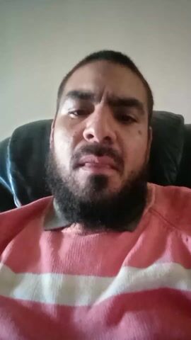 A man with a beard is sitting in a dark green recliner. He is wearing a pink and white striped sweater. He is looking directly at the camera. He begins by speaking then smiles. The background is a plain, light-colored wall.