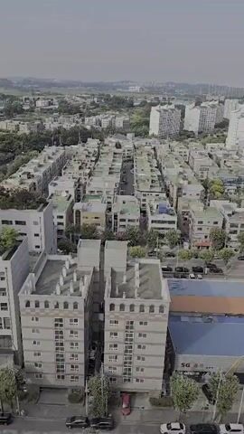 A drone shot pans over a dense urban landscape of apartment buildings and smaller residential structures. The buildings are mostly light-colored, with some featuring darker accents or blue roofs. Streets are visible between the buildings, with cars parked along the sides and some moving. In the foreground, there are several multi-story apartment buildings with visible windows and balconies. In the midground and background, the cityscape extends to a horizon marked by more apartment complexes and some greenery. The sky is overcast. A sign on one of the buildings in the midground reads "SG 스크린골프" and appears to be a business. Another building in the foreground has Korean text on its side that is partially visible and reads "지혜 TOWER". The overall impression is of a typical suburban or urban residential area.