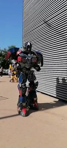 The video shows two individuals in elaborate robot costumes, resembling Optimus Prime and Bumblebee from the Transformers franchise, standing on a paved surface next to a building with a textured, metallic facade. The first shot features the Optimus Prime costume, which is predominantly blue, red, and silver, with intricate details. The person inside the costume shifts slightly. The camera then cuts to the Bumblebee costume, which is primarily yellow and black, with rounded features mimicking a classic car. The person in the Bumblebee costume also makes slight movements, turning its head. The background features a clear blue sky, green trees, and other people in the distance, suggesting an outdoor event or public space. The costumes are detailed and appear to be of high quality, designed to resemble the characters closely.