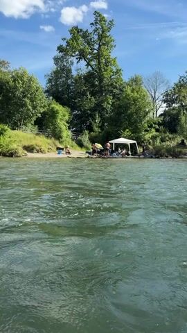 The video captures a sunny day by a river. In the foreground, the water is green and choppy, with white foam on the surface. The background features a sandy bank lined with trees. Several people are visible on the bank, some sitting and others standing. A white canopy tent is set up, with people gathered around it. One person walks into the water from the bank near the tent. The overall scene suggests a leisurely outing by the river.