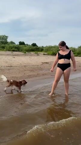 A young woman in a black bikini walks in shallow water on a sandy beach with a brown and white dog. The dog is in the water in front of her. In the distance, a person on a red paddleboard is visible in the water. The sky is cloudy.