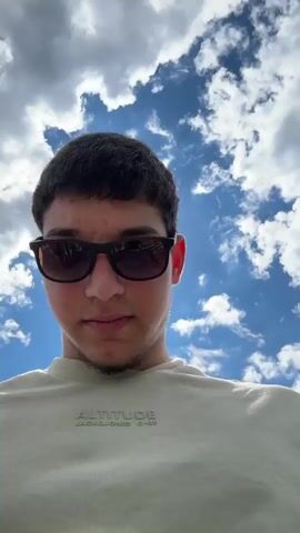 A young man with short dark hair and wearing sunglasses stands in front of a mountainous landscape. He is wearing a light-colored t-shirt with writing on it. The landscape is green, with a mix of trees and open areas. There is a town visible in the distance, nestled between the mountains. The sky is blue with puffy white clouds. The camera is at eye level and pans slightly to show more of the landscape. The man is looking directly at the camera for the first few seconds, then turns his head to admire the view. There is a black metal railing visible in the foreground.