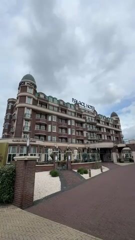 A large, ornate hotel building with multiple stories and a dark brick facade stands under a cloudy sky. The building features numerous windows, balconies, and two dome-like structures on its corners. Large signage on the roof clearly spells out "PALACE HOTEL" in white letters. To the left, a vertical sign is partially visible, reading "VAN DER VALK PALACE HOTEL NOORDWIJK," confirming the location. Another sign below it on the building appears to say "VLOED RESTAURANT."

In front of the hotel, a wide brick driveway leads towards the main entrance. On either side of the driveway, there are landscaped areas with white gravel and some green shrubbery, separated by low brick walls. A dark blue van is partially visible parked on the right side of the driveway. The overall impression is that of a grand and well-maintained hotel. No people are visible in the scene.