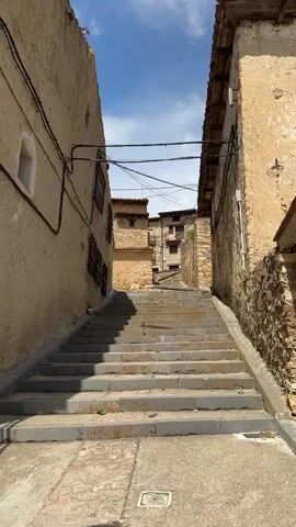 The video begins with a low-angle view of a narrow, cobblestone alleyway in a European village. The alley is flanked by tall, weathered buildings with stone and stucco exteriors. A set of stone stairs ascends the alley, casting long shadows in the bright sunlight. Overhead, utility wires are strung between the buildings. As the camera pans right, it reveals the corner of a building with large, rough-hewn stones integrated into its facade. A small, weathered wooden bench rests against the wall. Further to the right, a darker building with a metal grate over a window and a heavy wooden door is visible. A street sign is affixed to the wall above the door. The video continues to pan right, showing more of the building's facade and the street in front of it. The scene is quiet and appears to be uninhabited. The overall impression is one of historic charm and rustic simplicity.