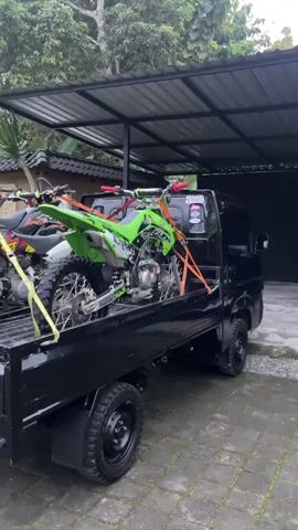 The video shows a black pickup truck parked under a covered structure. Two dirt bikes are secured in the truck bed using straps. The dirt bike closest to the camera is green, while the other is white and orange.  The background includes trees and a building with a dark door. The ground is paved.  There is no visible human interaction in the video.