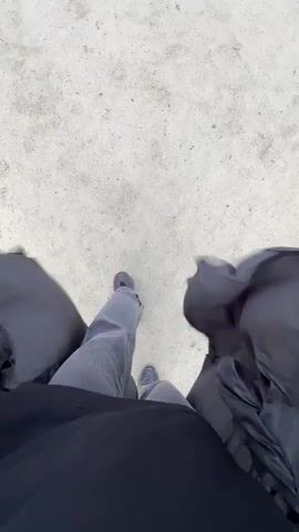 The video shows a first-person perspective of someone walking on a light-colored, uneven surface, possibly dirt or gravel. The person is wearing light-colored pants and dark-colored shoes. They are also wearing a long, dark, puffy jacket that is open and trails behind them, creating a sense of movement and possibly wind. The jacket obscures the lower body and sides of the person, directing the viewer's attention to their legs and feet as they walk. The lighting suggests it is daytime, but the overall atmosphere is somewhat muted. The camera is angled downwards, capturing the ground and the person's lower body. There are no other people or objects clearly visible in the frame. The sound is dominated by the rustling of the jacket and the sound of footsteps on the ground.