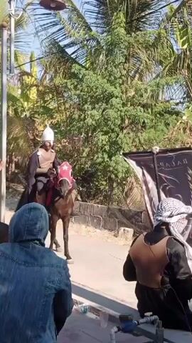A man in a helmet and a brown vest rides a brown horse with a red blanket on its head. He holds a black flag with white writing that says "BAL". People are watching him. The man is on a street with trees and palm trees in the background.