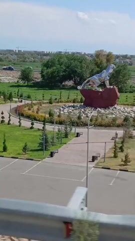 The video shows a large, metallic sculpture of a horse on a red, rocky base. The sculpture is located in a park-like setting with green grass, trees, and paved pathways. There are also benches and parking spaces visible in the foreground. The background consists of trees, buildings, and a slightly hazy sky. The camera appears to be handheld and is slowly panning and zooming out.