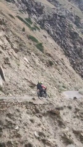 A motorcycle is seen riding on a narrow dirt road that snakes its way up a steep, rocky mountainside. The road is precarious, with a sheer drop on one side. Sparse vegetation clings to the rocky slopes, and the sky is overcast. Two people are riding the motorcycle, one driving and the other a passenger. The motorcycle is dark in color, and the riders are wearing helmets and protective gear. The scene appears to be in a remote, high-altitude region.