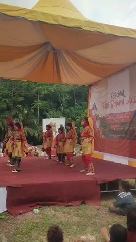 This is a vertical, first-person perspective video of a traditional dance performance taking place outdoors under a yellow and orange tent. A group of approximately seven young girls, dressed in colorful traditional costumes including gold tunics, red pants, and yellow headscarves, are performing a choreographed dance on a raised red stage. They are arranged in a line and appear to be synchronized in their movements. A banner with Indonesian text, likely related to the festival, is visible on the right side of the stage. In the foreground, the backs of several audience members are visible, seated on the grass. Behind the stage, a lush green forest can be seen. The lighting suggests it is daytime.