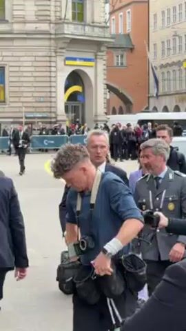 The video depicts a bustling scene in what appears to be a European city, possibly Munich, based on architectural styles and signage. The setting seems to be an outdoor public area, possibly a plaza or square. A group of people, including political figures, military personnel in uniform, photographers, and civilians, are walking together. The crowd includes a man who seems to be Jens Stoltenberg, Secretary General of NATO, based on his resemblance to publicly available images. Several men in suits, presumably security details, surround him. The presence of individuals in military attire with medals and decorations further suggests the importance of the event.  The backdrop includes buildings with visible windows, some adorned with the Ukrainian flag.  There is a sense of purpose and movement, indicating that the group is en route to a destination, likely related to a political or security event.