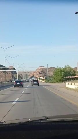 This media file is a video recording from inside a vehicle, likely a car, driving on a highway. The view is through the windshield, showing the road ahead. The highway has multiple lanes and is bordered by a concrete barrier on the left and a yellow guardrail on the right. 

**Subjects:**
*   **Objects:** Several cars are visible on the highway, including a black sedan in the foreground and a black SUV behind it, both in the right lane. Other vehicles are further down the road. Streetlights are positioned along the sides of the highway.
*   **Scene/Setting:** The scene is a highway passing through a mountainous or hilly area. The terrain is arid and rocky, with brown and tan-colored hills. There are some sparse green bushes and trees on the right side of the road. The sky is clear and blue, indicating good weather.

**Activity/Event:**
The primary activity is driving on a highway. The video captures the perspective of a driver or passenger as they travel along the road.

**Notable Details:**
*   **Time of Day:** The lighting suggests it is daytime, likely late morning or afternoon, given the clear blue sky and the angle of the sun casting shadows.
*   **Weather:** The weather appears to be clear and sunny.
*   **Location Cues:** The arid, rocky hills are characteristic of desert or semi-desert landscapes. The architecture of the streetlights and barriers might offer more specific regional clues, but without further context or clearer views, it's difficult to pinpoint a precise location beyond the general description. Given the provided location context of Qonaev, Kazakhstan, this landscape is consistent with the terrain found in that region.
*   **Emotions:** No explicit emotions are conveyed, as the video focuses on the driving environment.

**Visible Text:**
License plates are visible on some of the vehicles, but the text is too blurry to be read clearly.