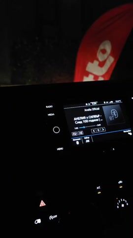 The scene shows a dark interior of a vehicle, likely a car, with the infotainment system illuminated. The screen displays music information, showing the artist "Anelia Official" and the song "след 100 години (Sled 100 godini)," playing via Bluetooth. The hazard lights button is lit, and the A/C and fan controls are also visible. Through the windshield, a blurred red banner with a white logo is partially visible, indicating an external element in the environment. The overall lighting is low, focusing on the dashboard components.