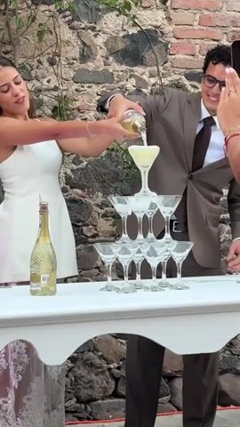 A bride and groom pour champagne into a glass tower on a white table at an outdoor wedding. The bride is wearing a short white dress and the groom is wearing a brown suit. A person off-screen to the right is recording the event with a cell phone. The tower is made of about 10 martini glasses. A champagne bottle sits on the table next to the tower. The background is a stone wall.